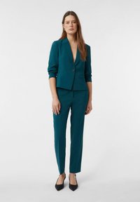 Woman wearing a teal tailored blazer and matching slim-fit trousers with black pointed-toe heels, standing against a plain white background.