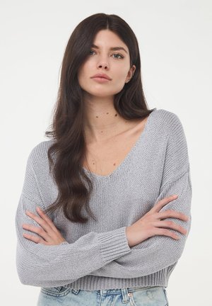 Maglione grigio a maglia con scollatura a V profonda, vestibilità ampia e polsini a coste. Indossato sopra jeans azzurri chiari con un motivo testurizzato.
