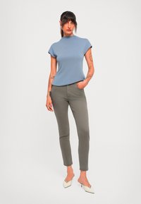 Kvinne med tatoveringer iført en blå t-skjorte med korte ermer, grå slim-fit bukser og hvite hæler som står mot en ensfarget hvit bakgrunn.