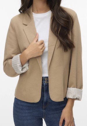 Vrouw draagt een beige blazer met opgerolde gestreepte manchetten, wit overhemd en blauwe spijkerbroek, houdt met één hand het revers van de blazer vast.