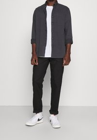 Camisa de botones de tela gris oscuro, combinada con una camiseta blanca, pantalones negros y zapatillas blancas con acentos negros.
