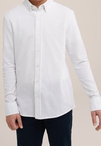 Ung person klädd i en vit knäppskjorta med långa ärmar och mörka byxor, stående med avslappnade armar längs sidorna mot en neutral bakgrund.