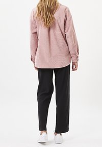 Camicia in corduroy rosa con maniche lunghe e polsini abbottonati, abbinata a pantaloni neri a gamba larga e sneakers bianche. Stile minimalista, vestibilità rilassata.