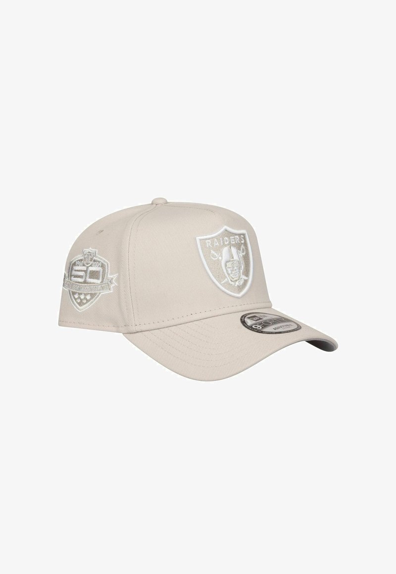 Beige Baseballcap mit gebogenem Schirm, ausgestattet mit weißen gestickten Logos, darunter das Raiders-Schild und ein Gedenkpatch.