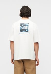 Άτομο με κοντά σκούρα μαλλιά που φοράει λευκό t-shirt με μπλε λογότυπο The North Face και κείμενο "Never Stop Exploring" στην πλάτη.