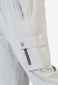 Lichtgrijze cargobroek met een zijzak, voorzien van een ritssluitingen en een spinnenlogo op de zakklep. Gemaakt van zacht, duurzaam materiaal.