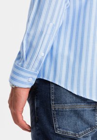 Camisa de rayas azul claro con puños abotonados, hecha de tela, combinada con jeans de mezclilla oscuros. Costuras visibles y un ajuste casual.