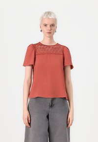 Top corto color ruggine con dettagli in pizzo ricamato sullo scollo e una vestibilità rilassata, abbinato a jeans grigi a vita alta.