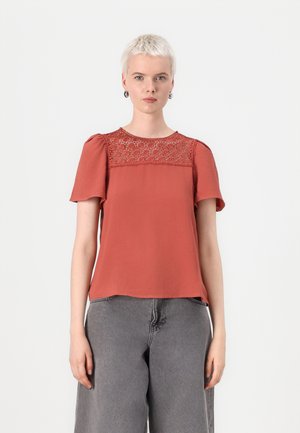 Rust-colored short-sleeve top with embroidered lace detail at the yoke and a relaxed fit, paired with gray high-waisted jeans.