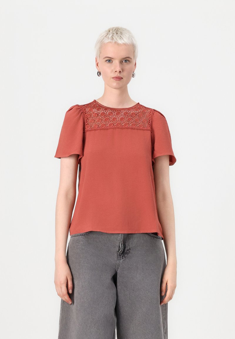 Top corto color ruggine con dettagli in pizzo ricamato sullo scollo e una vestibilità rilassata, abbinato a jeans grigi a vita alta.