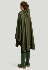 Oliivivihreä vedenpitävä sadetakki, jossa on huppu, löysä muotoilu, teksturoitu pinta ja heijastava yksityiskohta helmassa. Yhdistetty vihreisiin saappaisiin.