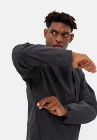 Black, waterproof jacket with a zippered side pocket. Features a high collar, adjustable cuffs, and smooth, matte texture. Casual fit.