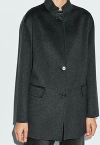 Cappotto di lana grigio scuro con colletto alla cinese, dotato di due tasche frontali e chiusura con un bottone. Tessuto liscio e vestibilità su misura.