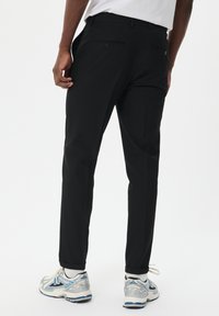 Pantalones negros de tela suave, con un corte ajustado, bolsillos laterales y bolsillos traseros con botón, combinados con zapatillas deportivas grises.