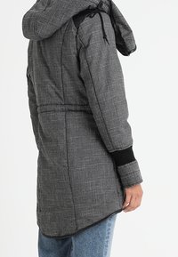 Gråmönstrad parkajacka med huva, dragsko, elastiska ärmslut och smalnad midja. Har en hållbar tygtextur och en längre bakre f åll.