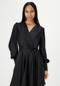 Robe noire ajustée en tissu lisse. Elle possède de longues manches bouffantes, une taille cintrée avec une ceinture nouée et une jupe fluide.