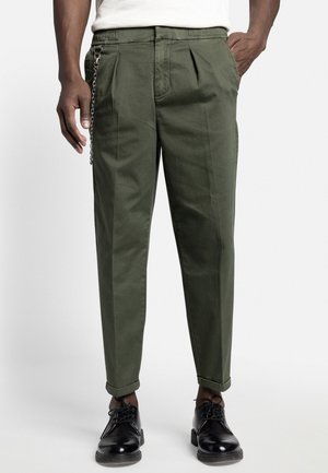 Homme portant un pantalon plissé vert olive avec des revers retroussés, des chaussures habillées en cuir noir, une chemise blanche et une chaîne en métal suspendue à la poche.
