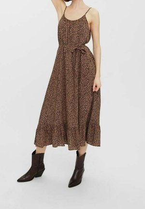 Femme portant une robe midi en imprimé léopard brun avec des fines bretelles et une ceinture à nouer, associée à des bottines marron foncé, se tenant sur un fond blanc.