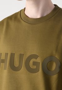 Gros plan d'une personne portant un sweat-shirt vert olive à col rond avec de grandes lettres mates "HUGO" sur la poitrine.