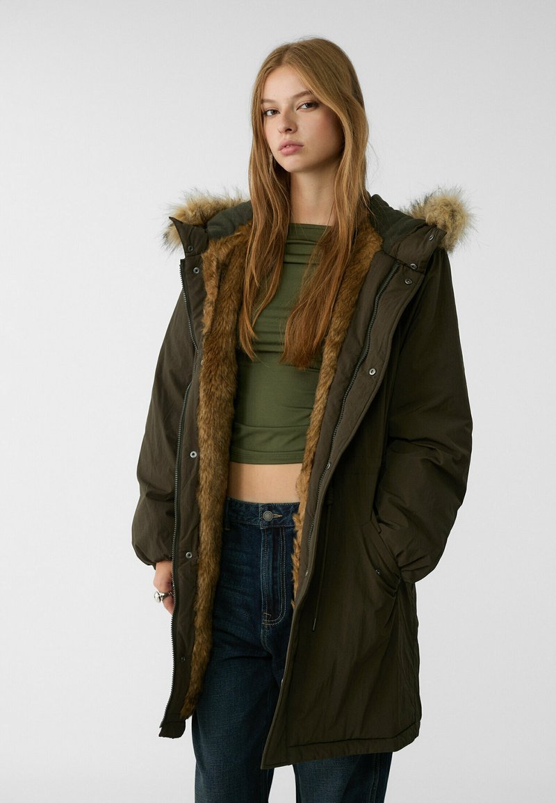 Parka olive avec doublure en fausse fourrure et capuche, dotée d'une fermeture éclair et de boutons-pression, ainsi que de poches latérales. Portée sur un haut vert et un jean.