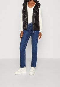 Black puffer vest with a hood over a white long-sleeve top, paired with dark blue jeans and white sneakers. Smooth, shiny texture.