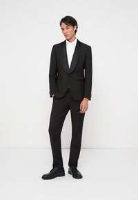 Jeune homme se tenant contre un mur blanc, portant un smoking noir avec des revers en satin, une chemise blanche et des chaussures de ville noires.