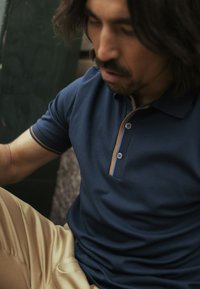 Laivastonsininen poolopaita, jossa on vastavärinen beige kaulus, kolme nappia ja istuva malli, yhdistettynä beigenvärisiin housuihin. Sileä kangaspinta.