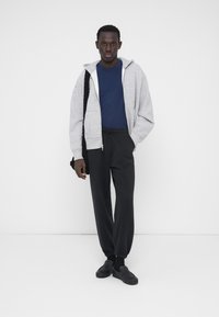 Sudadera gris con cremallera, camiseta azul, pantalones de chándal negros y zapatos negros sin cordones. La sudadera tiene un bolsillo frontal y capucha con cordón.