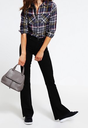 Donna che indossa una camicia a quadri e pantaloni neri svasati, con una borsa grigia in mano, in piedi con una gamba leggermente sollevata su sfondo bianco.