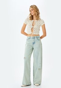 Virkat krämfärgad topp med kort ärm, diamantmönster och framsida med snörning, kombinerad med ljusblå, vida, slitna jeans och krämfärgade skor.