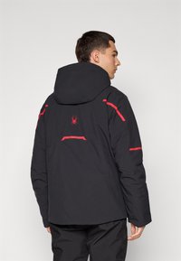 Jachetă impermeabilă neagră cu glugă. Are accente roșii și un logo cu păianjen pe spate. Design elegant cu o textură netedă.