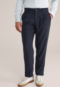 Man in een donkerblauwe op maat gemaakte broek, lichtblauw shirt en witte sneakers met rubberen zolen tegen een neutrale achtergrond.