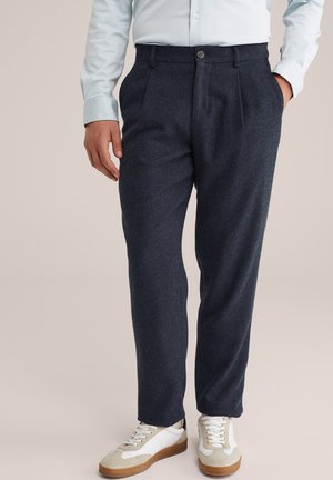 Man in een donkerblauwe op maat gemaakte broek, lichtblauw shirt en witte sneakers met rubberen zolen tegen een neutrale achtergrond.
