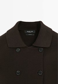 Brauner gestrickter Baumwollcardigan mit breitem Kragen und zwei schwarzen Knöpfen. Das Etikett trägt die Aufschrift "Massimo Dutti" mit Pflegehinweisen darunter.