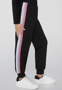 Schwarze Jogginghose mit seitlichen Streifen in Pink, Hellblau und Silber. Mit elastischem Bund und Bündchen. Kombiniert mit weißen Sneakers.