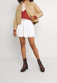 Beżowa kurtka z sztucznej skóry, czerwona dzianinowa koszulka, białe plisowane szorty i brązowe botki na obcasie. Prosty fason, teksturowane materiały.