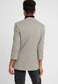 Kostkovaný blazer v béžové a černé barvě, který má strukturovaný design, rukávy po tři čtvrtiny a kontrastní černý límec. Texturovaná tkanina.