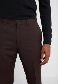 Uomo che indossa pantaloni tailored marroni scuri e una maglietta nera a maniche lunghe, con la mano destra poggiata accanto a lui su uno sfondo bianco.