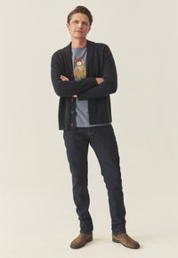 Tumšs džemperis ar ribotiem elementiem, valkāts pār zilu grafikā t-kreklu ar laci. Apvienots ar tumšām džinsēm un brūnām zamšādas zābakiem.