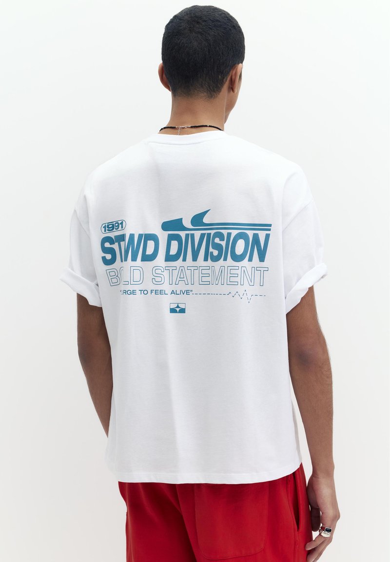 Homme aux cheveux courts et foncés portant un T-shirt blanc avec un texte bleu « STWD DIVISION B.D. STATEMENT » et un pantalon rouge, debout de dos.