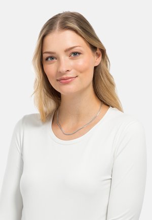Silberne Kettenhalskette mit einem wellenförmigen Design, getragen über einem weißen Langarmoberteil. Die Kette hat eine strukturierte Oberfläche und eine dezente reflektierende Qualität.