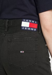 Vaqueros de denim negro con un parche visible de "TOMMY JEANS" en marino, rojo y blanco en la cintura y un pequeño logo en el bolsillo trasero.