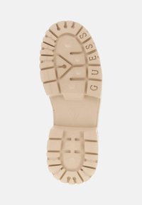 Talpa de cauciuc bej cu un model texturat, branding și șanțuri distincte pentru aderență. Prezintă logo-ul "GUESS" evidențiat pe o parte.