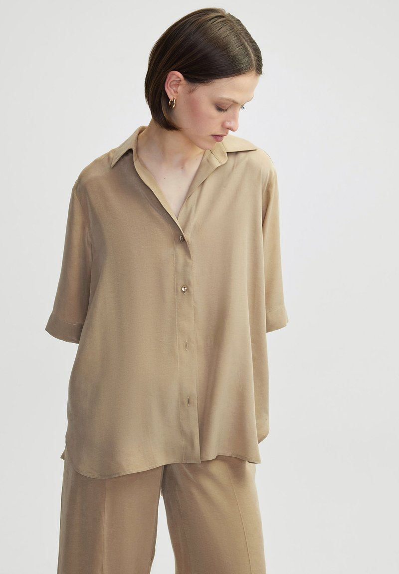 Tan, short-sleeve button-up shirt made of smooth fabric with a collar, featuring a relaxed fit and subtle sheen, complemented by matching pants.
