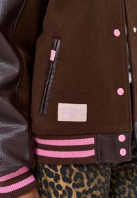 Zbliżenie brązowej kurtki varsity z różowymi guzikami, w paski mankiety, skórzany wykończenie kieszeni oraz naszywka z napisem "Bądź powodem, dla którego ktoś się uśmiecha dzisiaj."