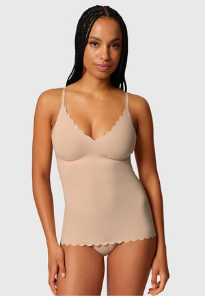 Mujer con el cabello trenzado, vistiendo una camiseta sin costuras color nude y ropa interior a juego con bordes ondulados, posando contra un fondo claro liso.
