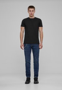 Fekete pamut póló, rövid ujjú, kék rongyos, slim-fit farmernadrággal és fekete sportcipővel, egy sima szürke háttér előtt állva.