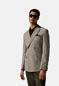 Blazer gris à double boutonnage en tissu texturé, avec des boutons dorés et un mouchoir de poche, associé à un polo noir et un pantalon rayé.