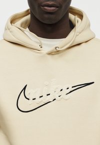 Bež bombažni pulover s kapuco, ki ima teksturiran bel napis "nike" in črni logotip Swoosh, s kapuco z zavezovanjem in mehko tkanino.