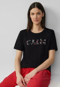 Zwarte katoenen t-shirt met korte mouwen, versierd met kleurrijke paillette applicaties die "CALM" op de borst spellen. Gecombineerd met rode broek.